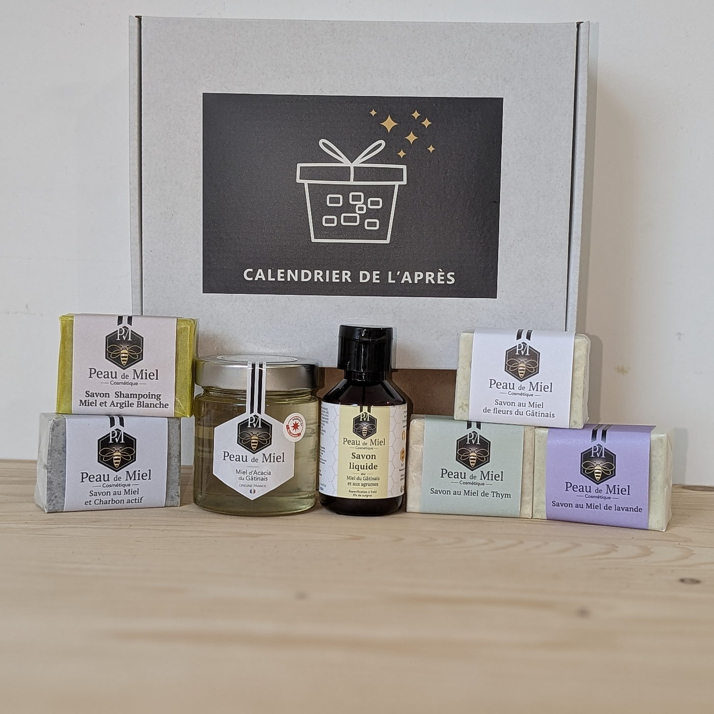 Ensemble des produits du Calendrier de l'Après, savons Peau de Miel dans leurs étuis, pot de miel et flacon ambré devant la boîte blanche